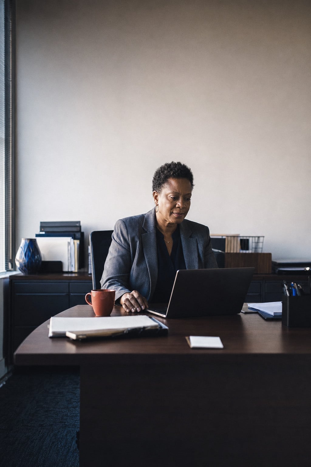 A manager at her desk works on a laptop, a cup and a folder beside her