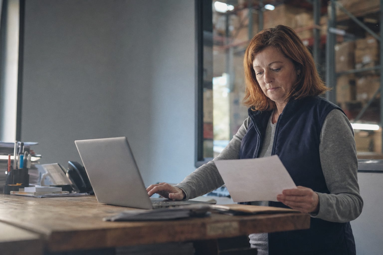 An operations coordinator enters an order from a printed sheet, the warehouse behind her