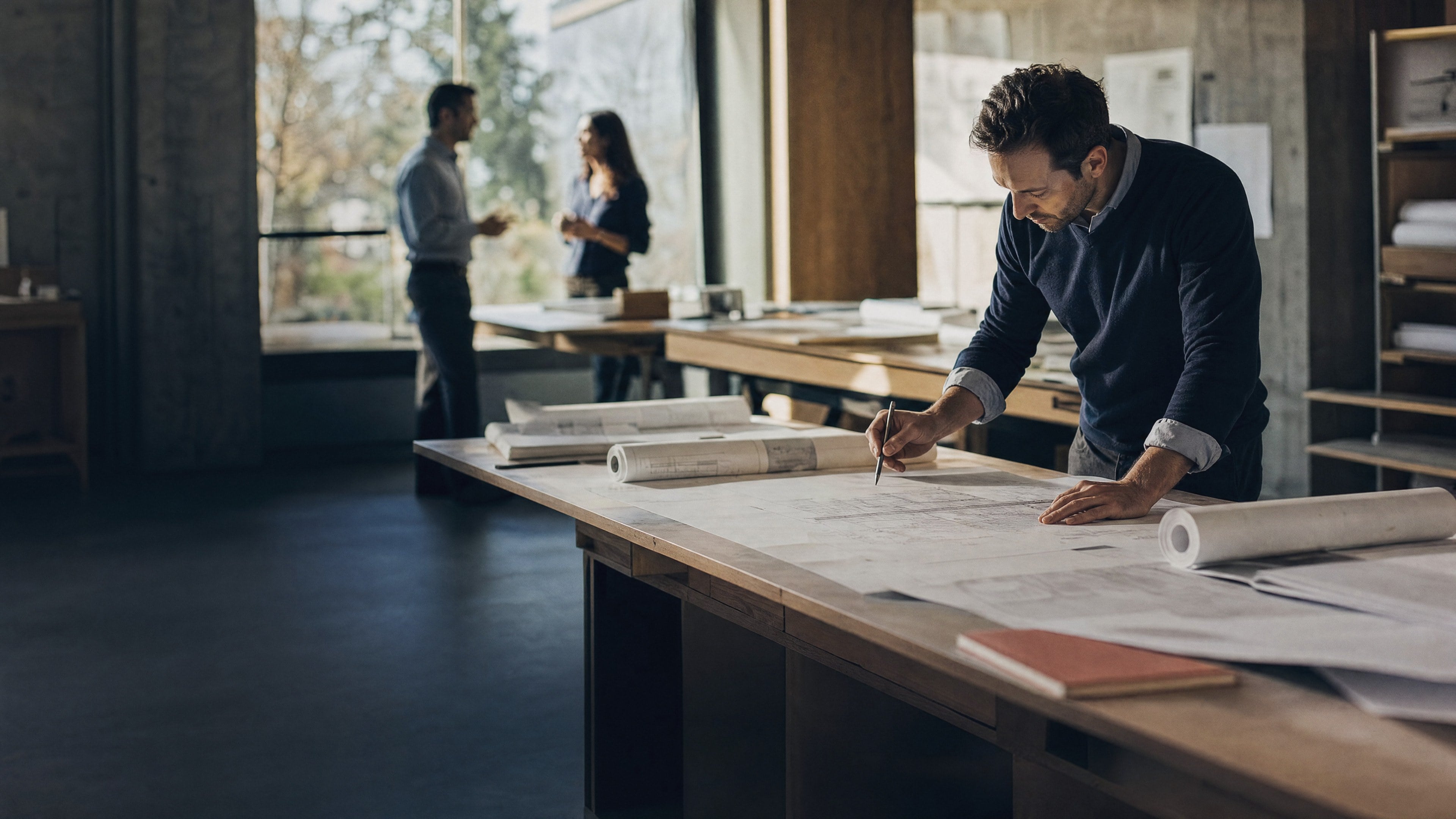 An engineer marks up a drawing on a long worktable while two colleagues talk by the window