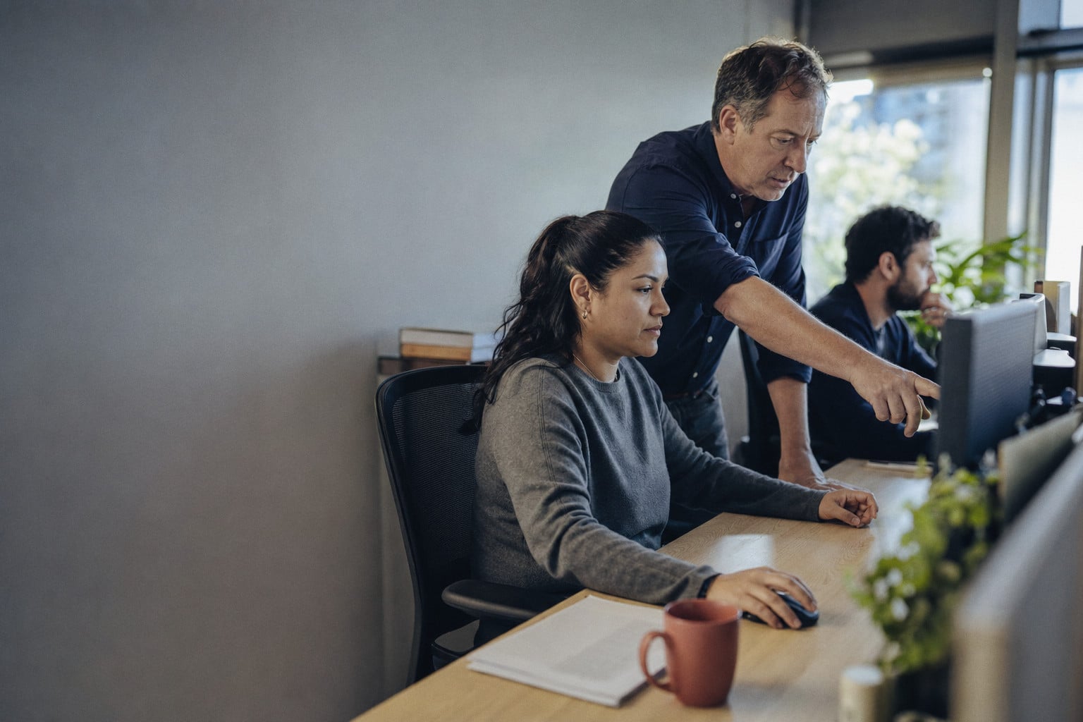 A woman works at her screen while a colleague leans in and points, a third person at the next desk